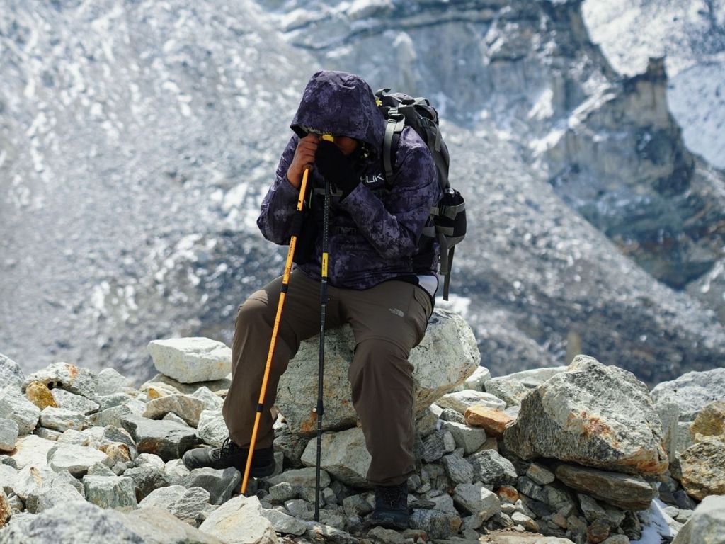 Breathing Easy at Altitude: Top 5 Reasons to Use a Concentrated Oxygen Machine 2 A hiker in a purple jacket sits on rocky terrain, resting with trekking poles, surrounded by a snowy mountain landscape.