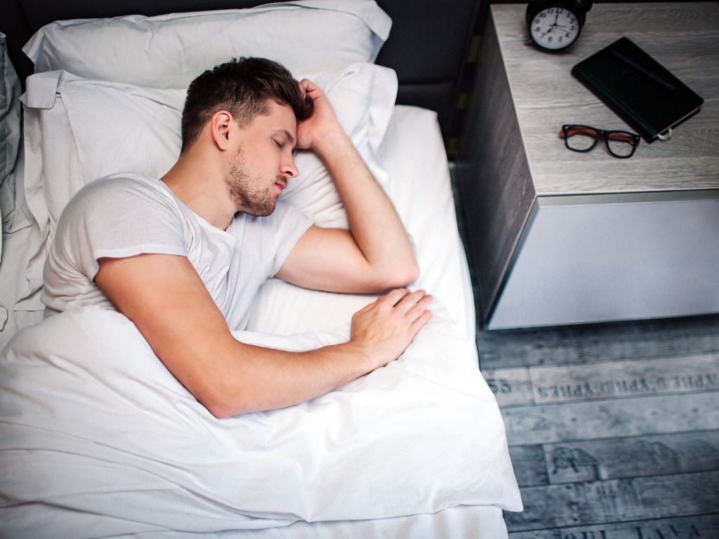 Breathing Easy at Altitude: Top 5 Reasons to Use a Concentrated Oxygen Machine 5 A young man sleeps on a cozy bed, surrounded by white bedding, with a clock and glasses on a nightstand beside him.